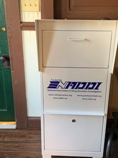 White secure drop-off box with National Association of Drug Diversion Investigators logo and website links, placed indoors.
